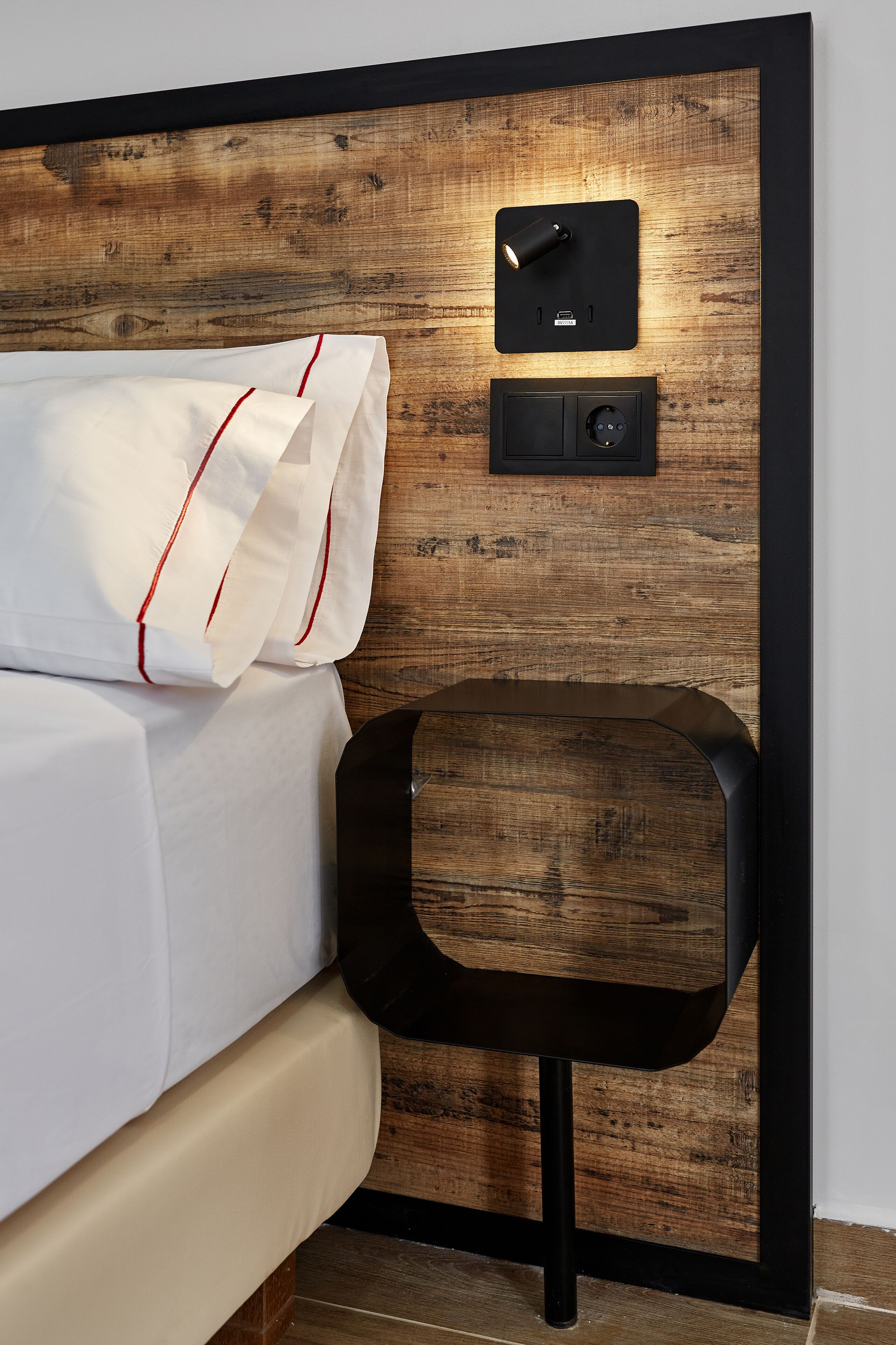 A neatly made bed with white linens and red piping, a bedside table with a black finish, and a wall-mounted control panel.