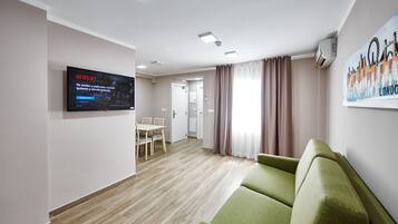 A modern living room with a green sofa, a flat-screen TV displaying Netflix, a dining area with a table and chairs, and a painting of London on the wall.