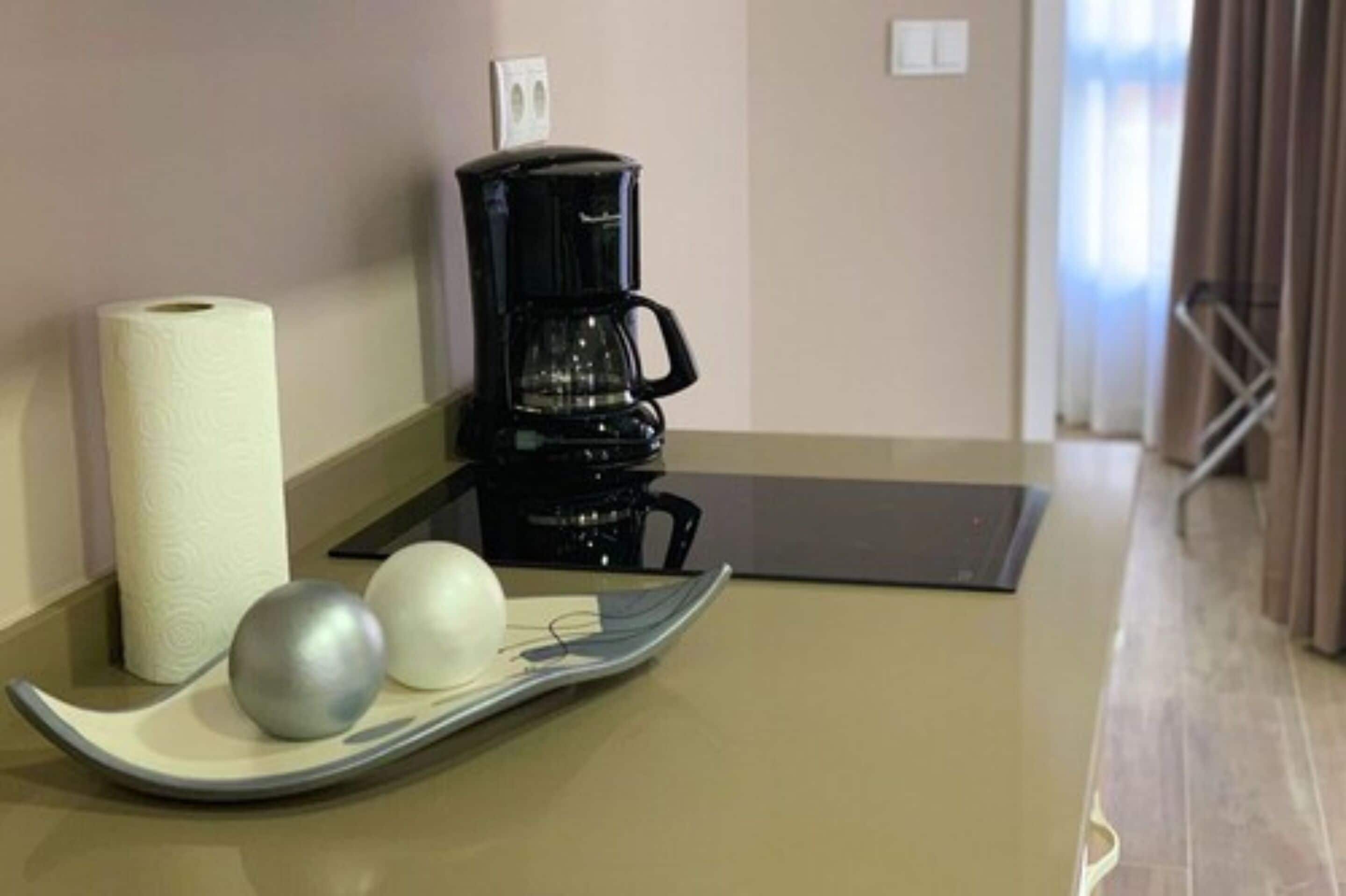 Una cafetera, un rollo de papel higiénico y dos objetos esféricos sobre una bandeja en una habitación con suelo y paredes color beige.