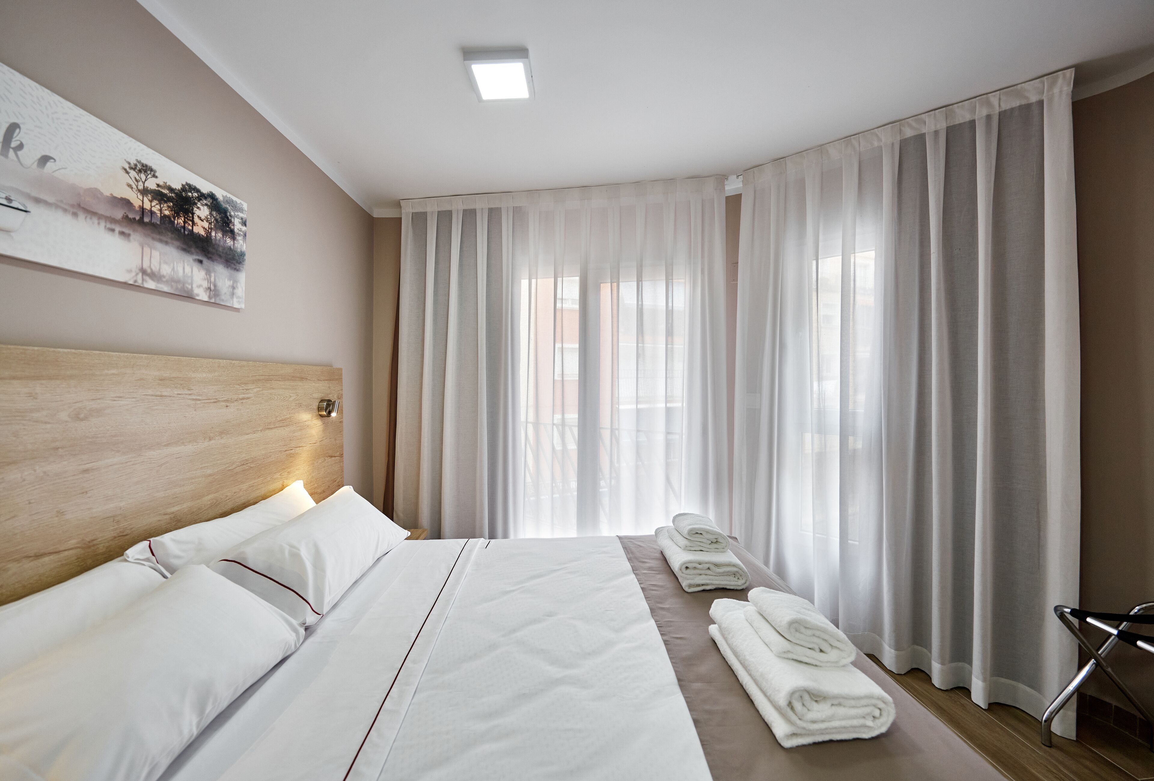 A hotel room with a large bed, a headboard, a painting on the wall, a window with sheer curtains, and a towel rack with towels.