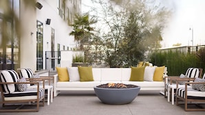 Un área de estar exterior moderna con un hogar, sofá blanco, sillas a rayas y un árbol grande al fondo.