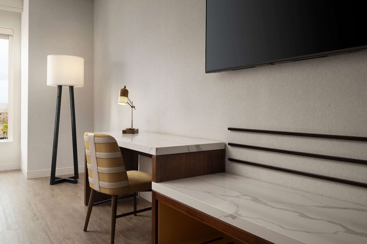 A modern hotel room with a flat-screen TV mounted on the wall, a desk with a chair, and a floor lamp.