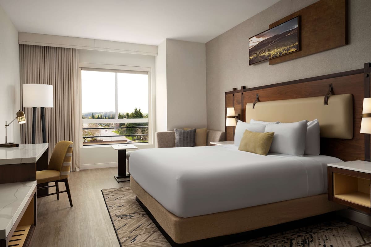 A hotel room with a large bed, a desk, a chair, a small table, and a window with a view.