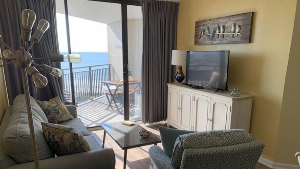 Una sala de estar con sofá, sillón, mesa de centro y televisor sobre un mueble bajo. Hay un balcón con mesa y sillas, y vista al mar.