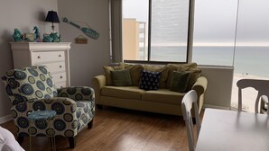 Una sala de estar con un sillón estampado, un sofá color beige con almohadas náuticas, una mesita y vistas a la playa a través de amplios ventanales.