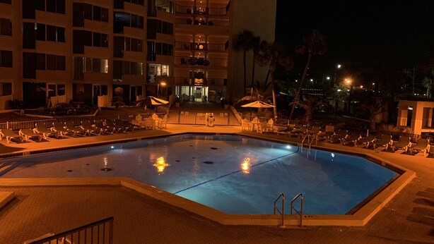 Piscina con sillones de descanso y sombrillas, rodeada por un edificio de varios pisos con balcones.