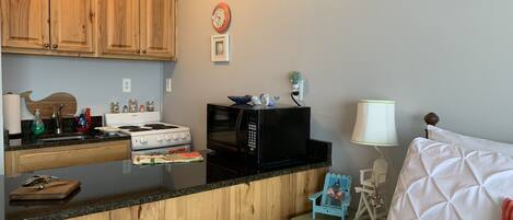 A compact kitchen with wooden cabinets, a microwave, a stove, and a small bedside table with a lamp.