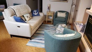 Un salon avec un canapé blanc, un fauteuil turquoise et un pouf, un tapis à rayures et une petite table d’appoint avec une lampe.