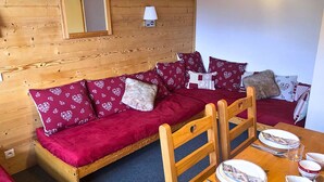 Un accogliente interno di una baita con un divano in legno, un tavolo da pranzo con piatti e posate, e una piccola lampada a parete.
