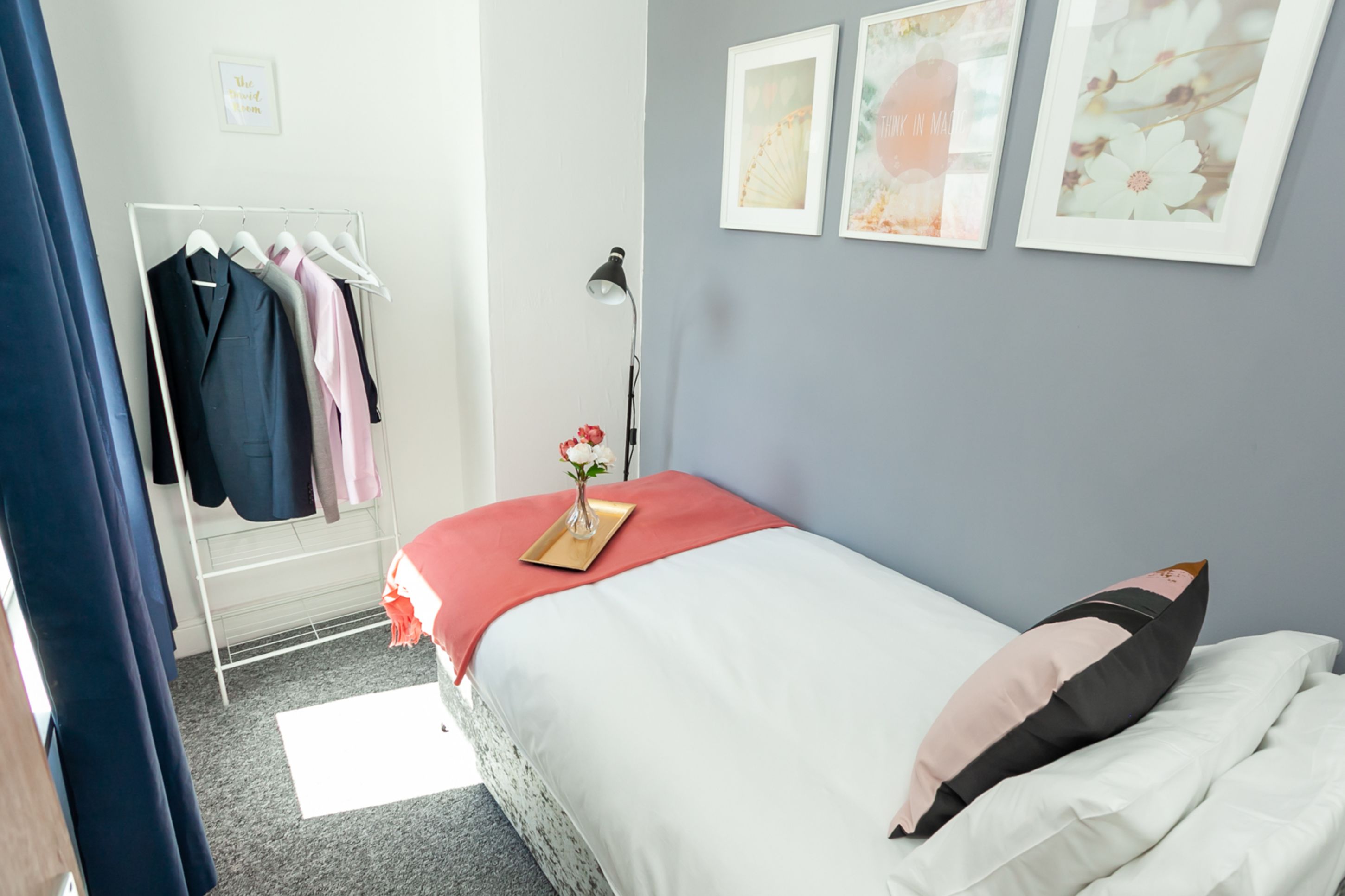 Un dormitorio con una cama, una mesita de noche con un florero, un perchero y cuadros en la pared.