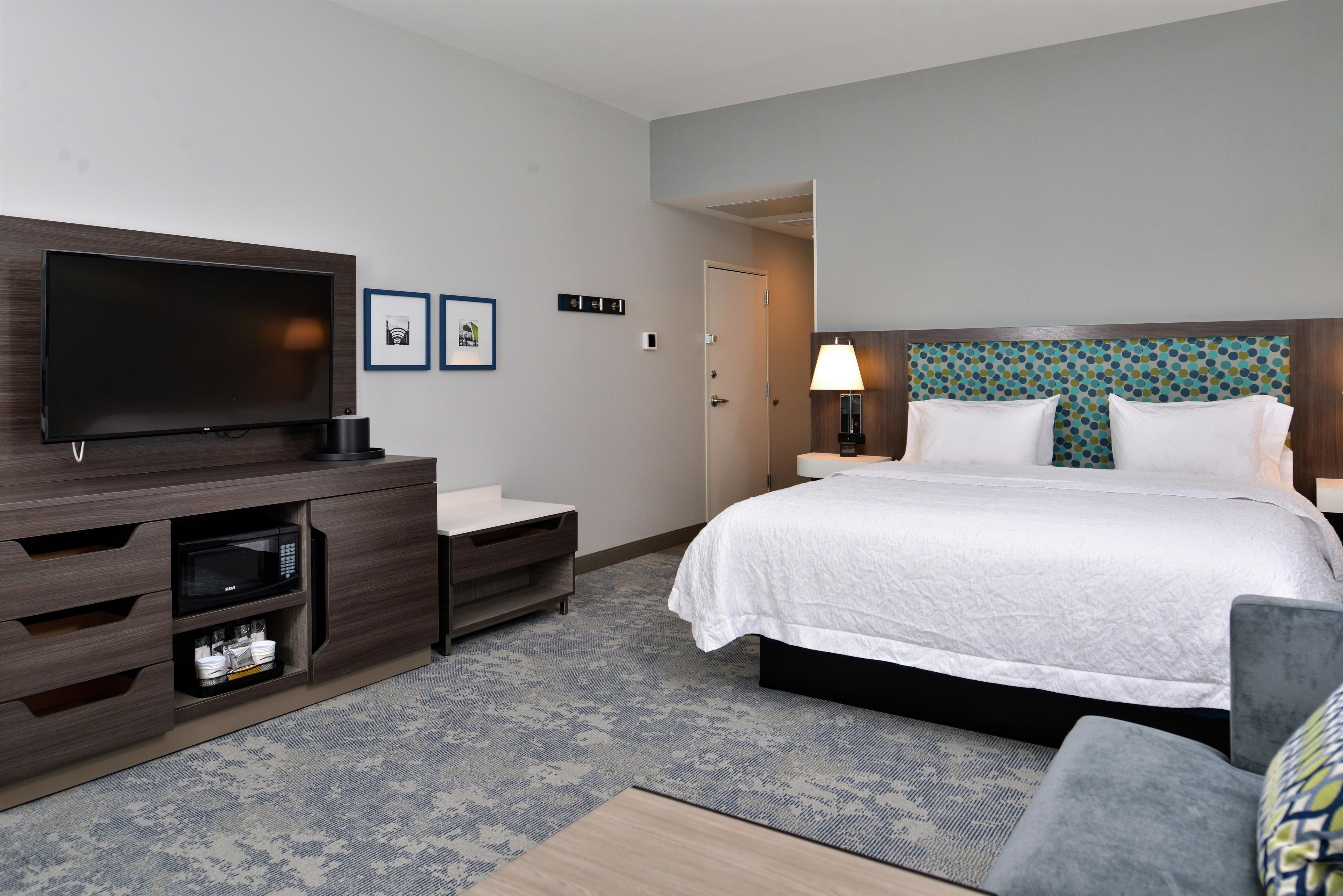 A hotel room with a flat-screen TV, a bed with white bedding, a desk with a lamp, and a patterned carpet.