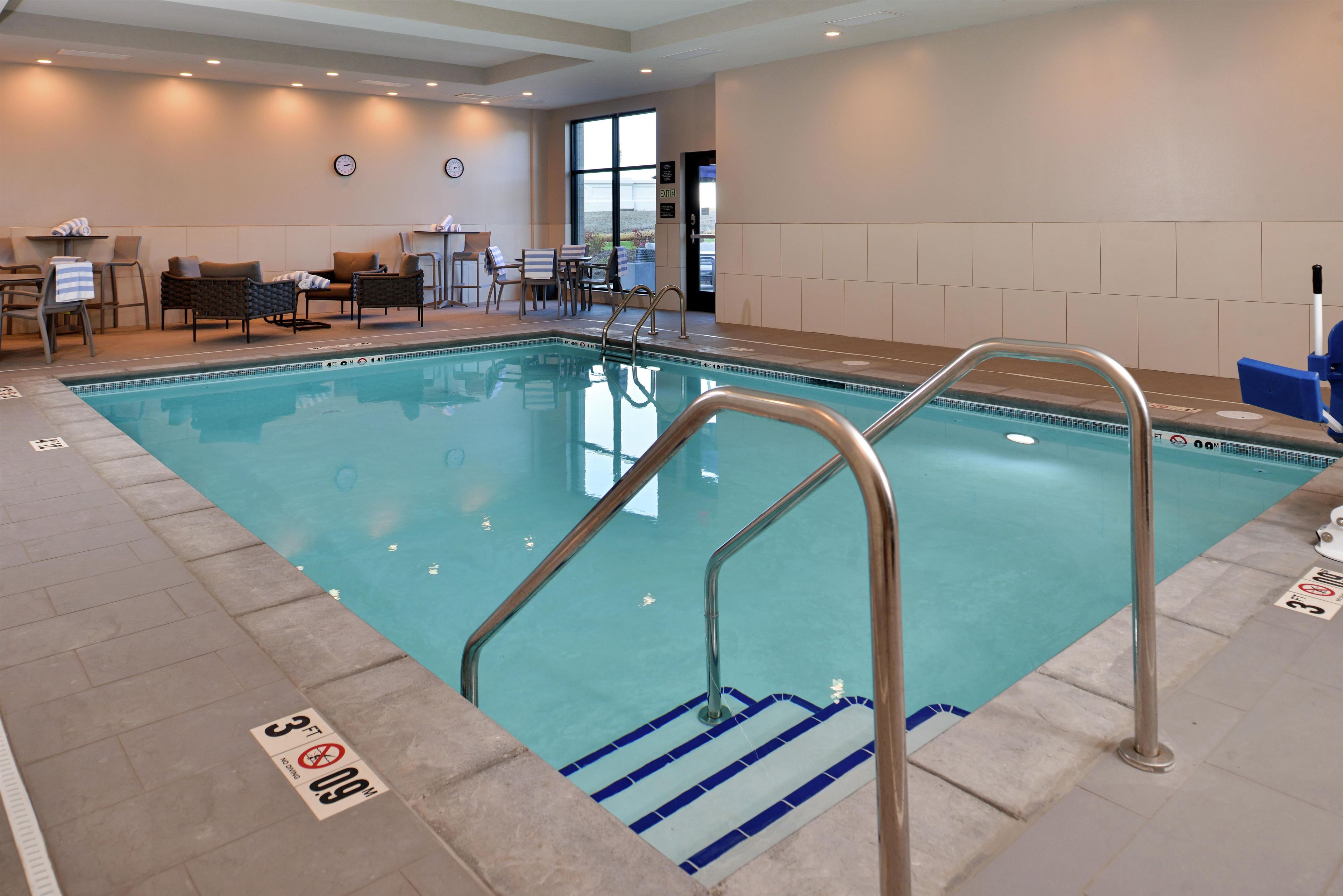 An indoor swimming pool with clear water, a metal ladder, and a seating area with chairs and tables.