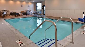 An indoor swimming pool with clear water, a metal ladder, and a seating area with chairs and tables.