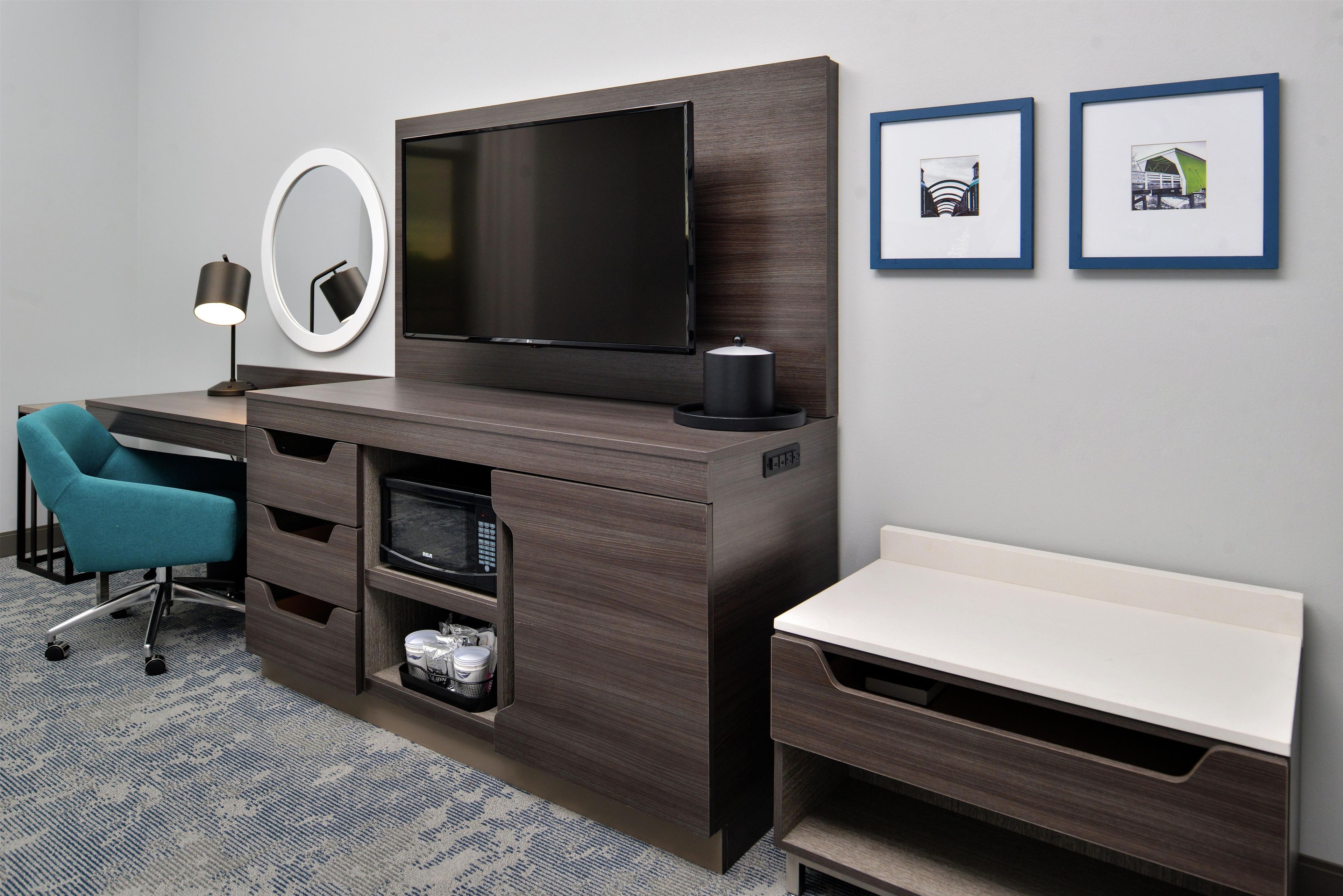 Une chambre d’hôtel équipée d’un téléviseur à écran plat, d’un bureau avec une chaise, d’un banc, d’un four à micro-ondes et d’un petit réfrigérateur. Deux tableaux encadrés ornent le mur.