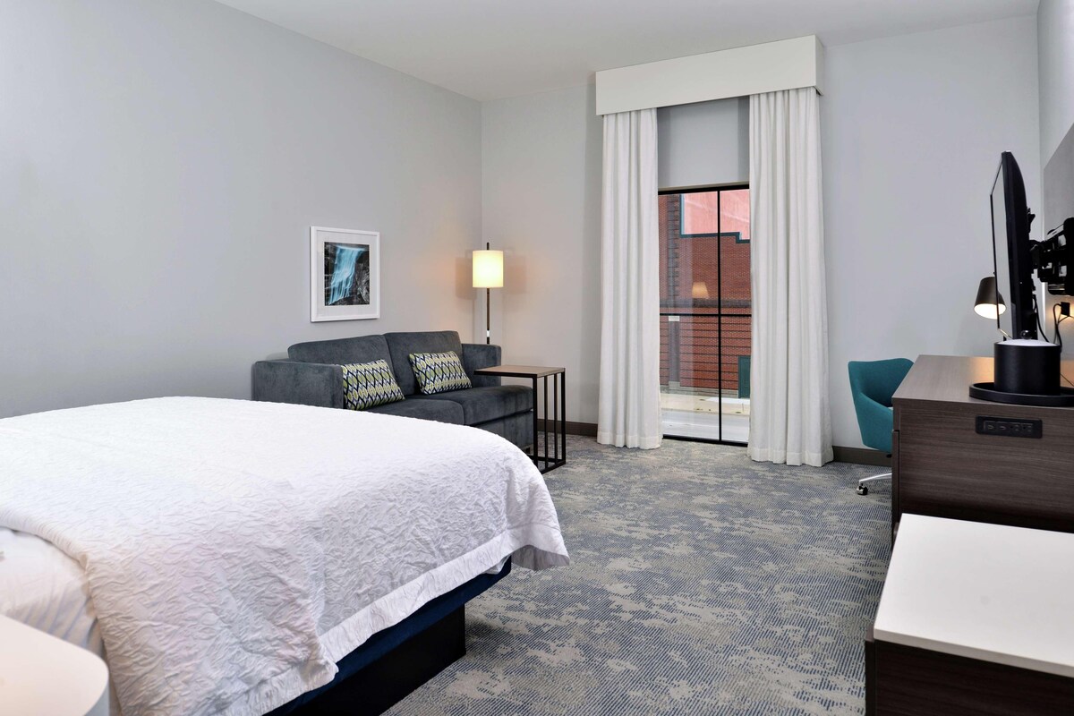 Habitación de hotel con una cama grande, un sofá, un escritorio, una lámpara y vistas a un edificio de ladrillo por la ventana.