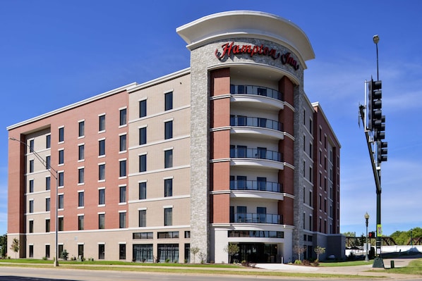A Hampton Inn hotel with a curved corner facade, multiple balconies, and a clear blue sky.