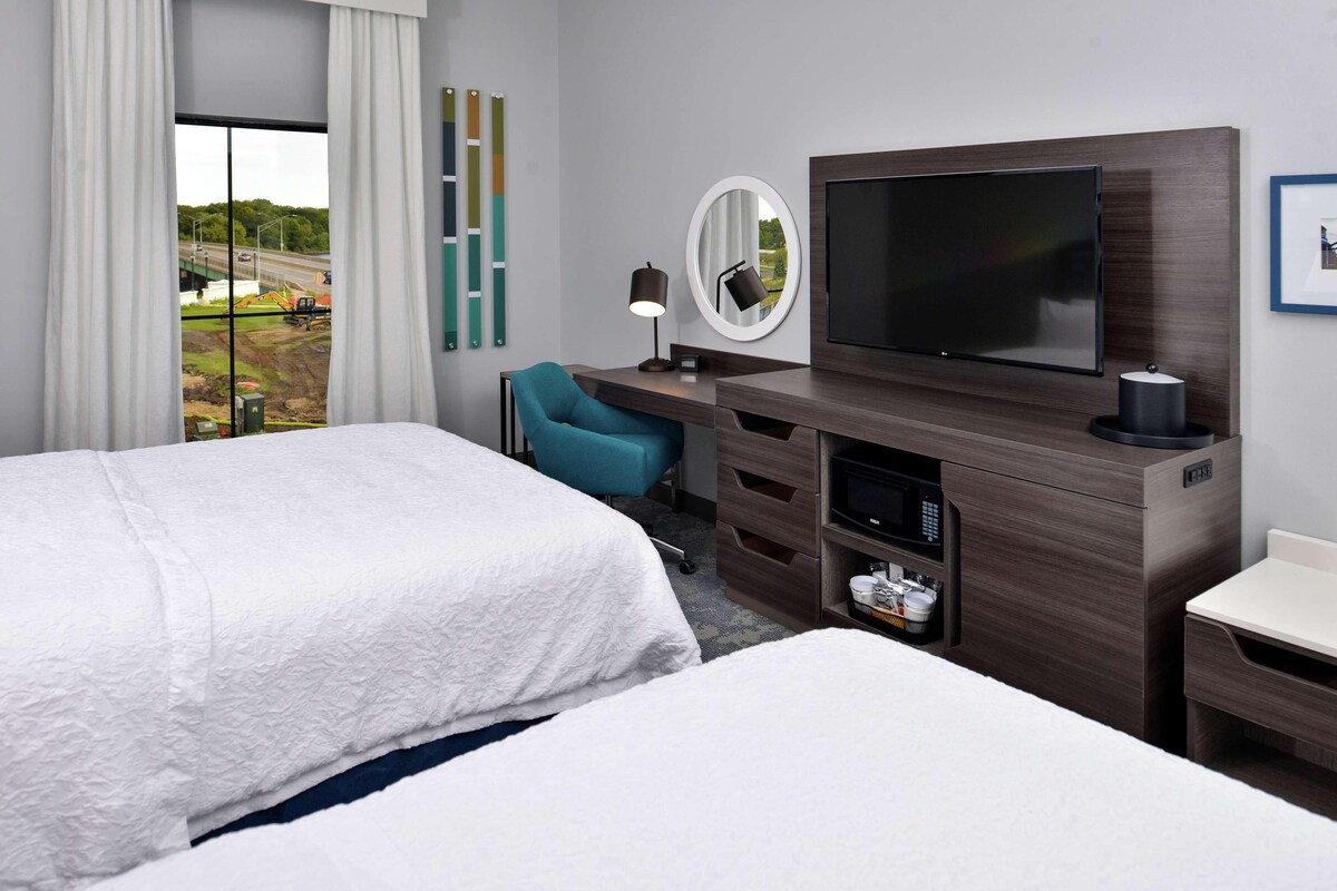 Habitación de hotel con dos camas, un escritorio con televisor, un espejo y vista a una obra en construcción.