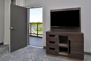 A room with a large flat-screen TV mounted on a wooden cabinet with drawers, a door leading to a balcony with a view of a river and greenery.
