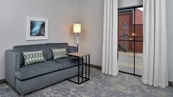 A hotel room with a grey sofa, patterned pillows, a small table, a floor lamp, and a view of a brick building through a window with white curtains.