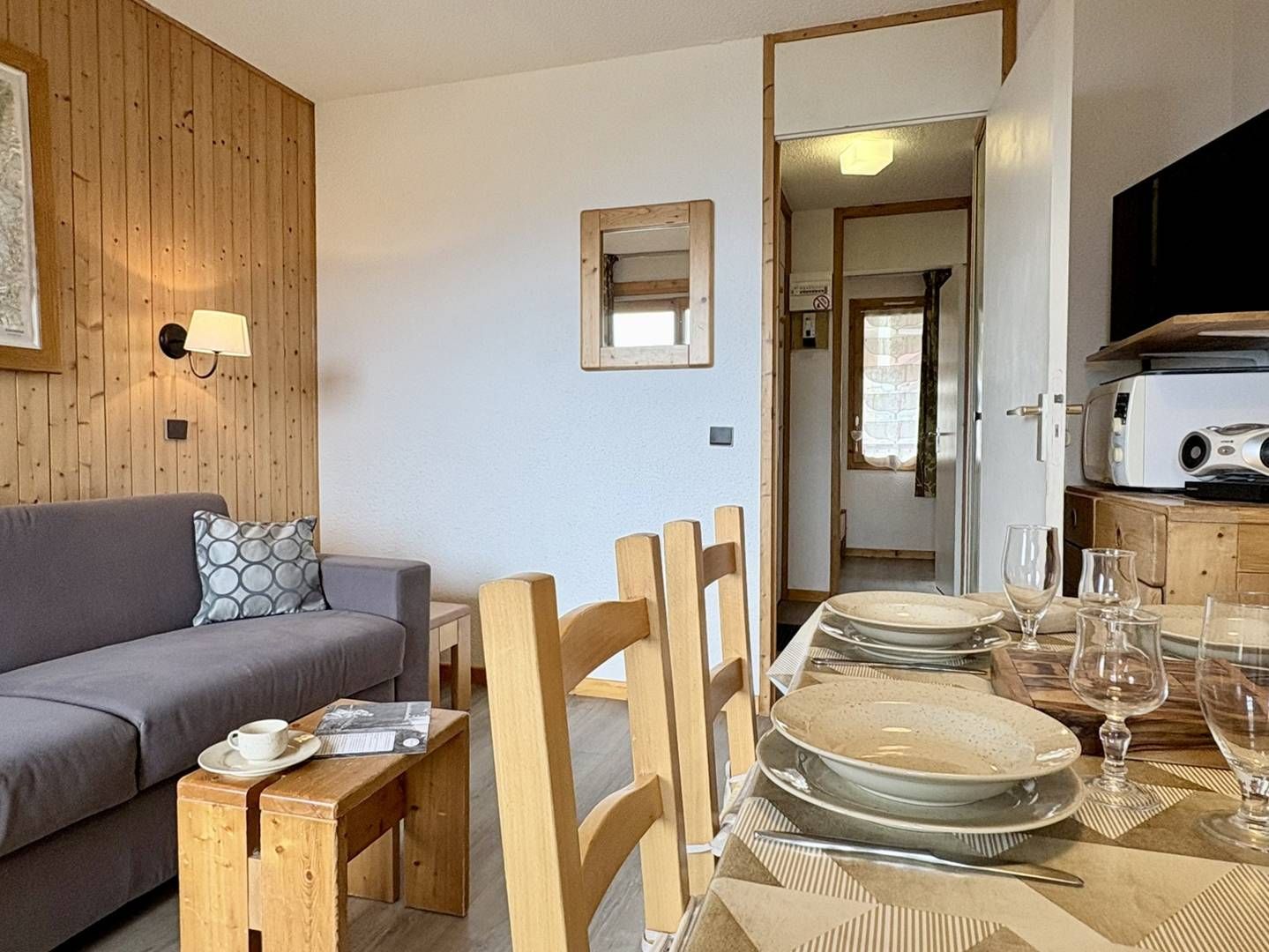 A cozy living area with a sofa, a dining table set with plates and glasses, wooden chairs, and a television on a wooden cabinet.