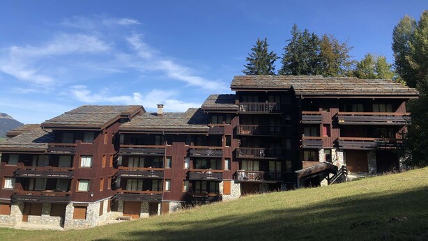 A multi-story building with balconies and a stone facade, surrounded by trees and a clear sky.