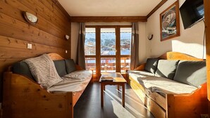A cozy wooden cabin with two wooden sofas, a small table, and a large window offering a view of a snowy mountain landscape.