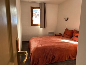 A bedroom with a large bed, a window with curtains, a bedside table, and a wall-mounted light fixture.
