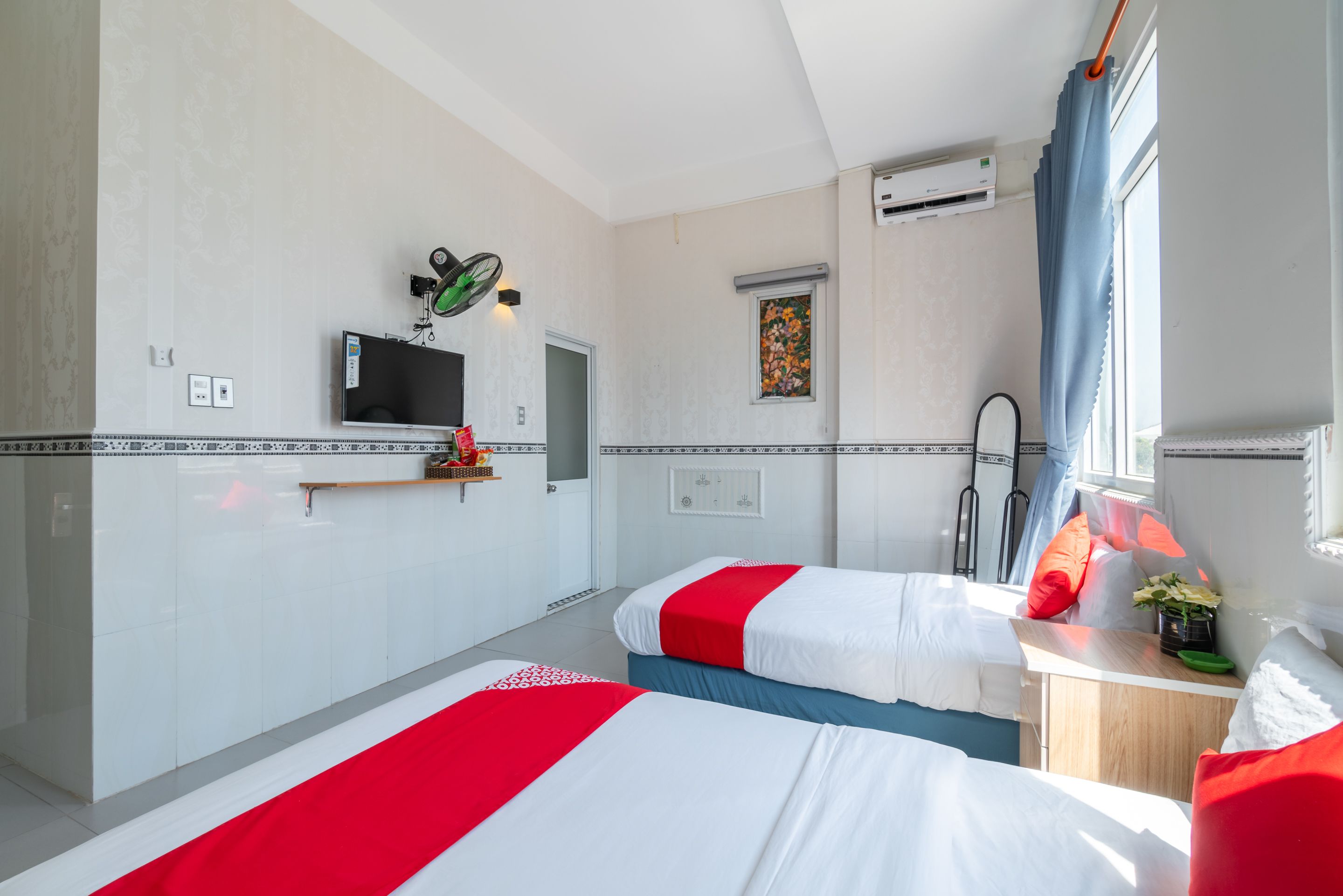 A hotel room with two beds, a TV mounted on the wall, a small desk with a plant, and a window with curtains.