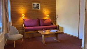 A cozy room with a wooden wall, a sofa with red cushions, a wooden coffee table, and two wall-mounted lamps. - Bûche 13 - Cosy studio with movable partition (LES AVANCHERS VALMOREL)