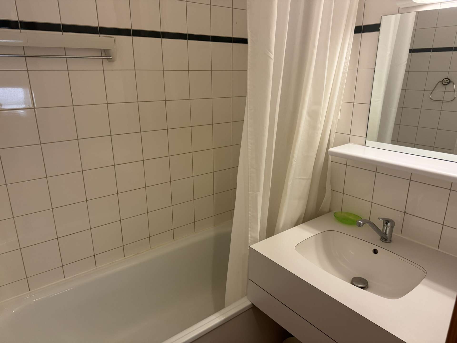 A bathroom with white tiled walls, a shower with a white curtain, a sink with a green soap dish, and a mirror above the sink.