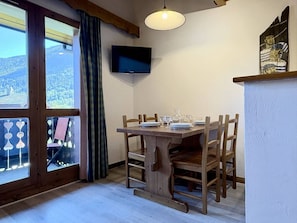 Ein Essbereich mit Tisch für vier Personen, Holzsesseln, einem an der Wand montierten Fernseher und Blick auf eine Berglandschaft durch große Fenster.