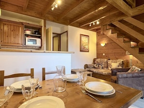 A wooden dining table set with plates, glasses, and cutlery, a sofa with patterned cushions, and a staircase with wooden railings.