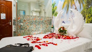 A hotel room with a bed adorned with rose petals and a bouquet, a wooden door, a shower area, and a tropical mural.