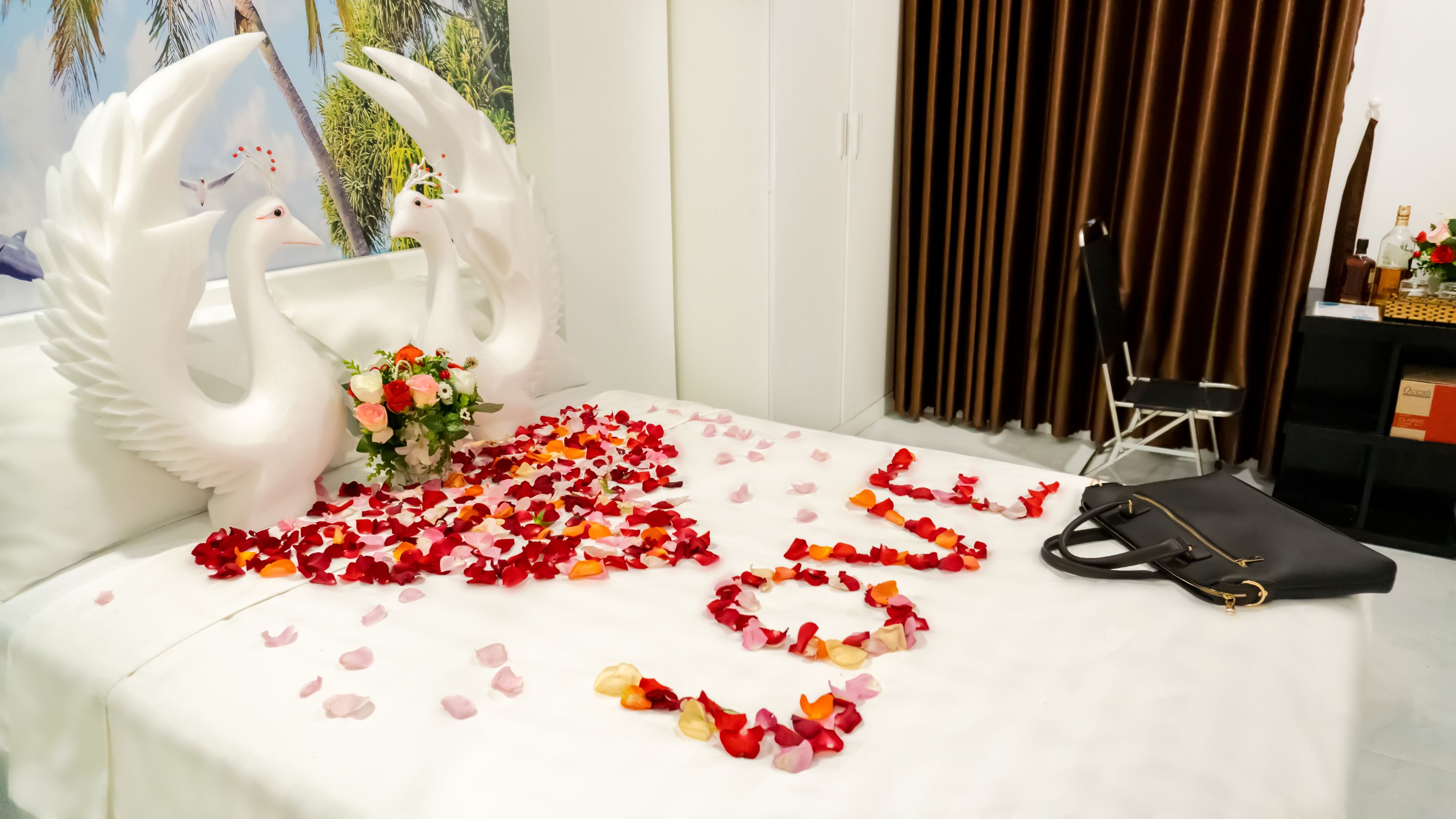 Un lit décoré de pétales de rose disposés en forme de cœur, un cygne blanc en guise de décoration, et un sac à main noir posé sur un couvre-lit blanc.
