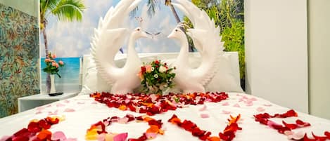 A bed with a white bedspread, two white swan-shaped pillows, a bouquet of flowers, and rose petals arranged on the bed.