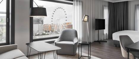 A modern hotel room with a large window, a chair, a bed, a desk, and a lamp.