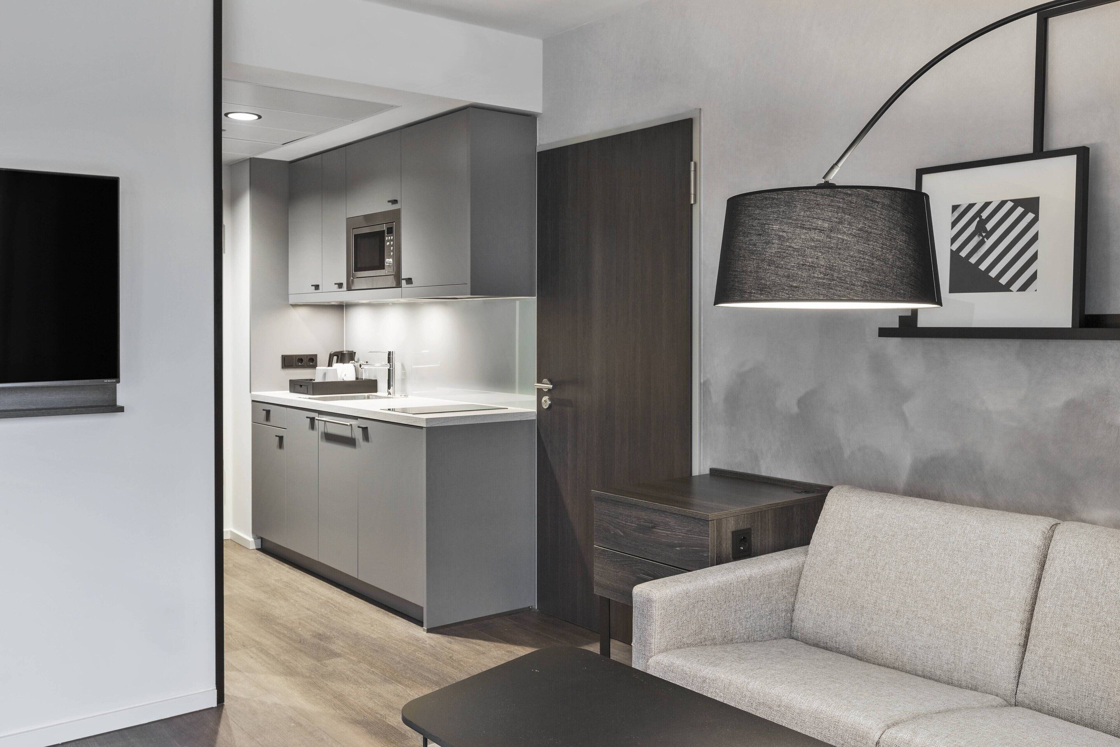 A modern kitchen with grey cabinets, a microwave, and a sink. There is a dark wooden door, a grey sofa, and a black table.