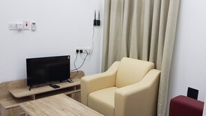 Una habitación con un televisor sobre un soporte de madera, un sofá color beige, una mesa de madera y un banco rojo.