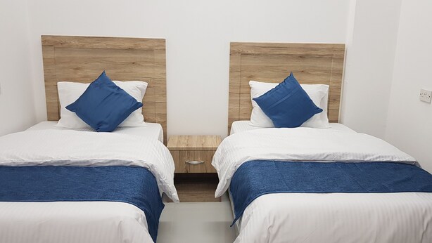 Habitación de hotel con dos camas, cada una con cabecero de madera, ropa de cama blanca y almohadas azules. Hay una mesita de noche de madera entre las camas.