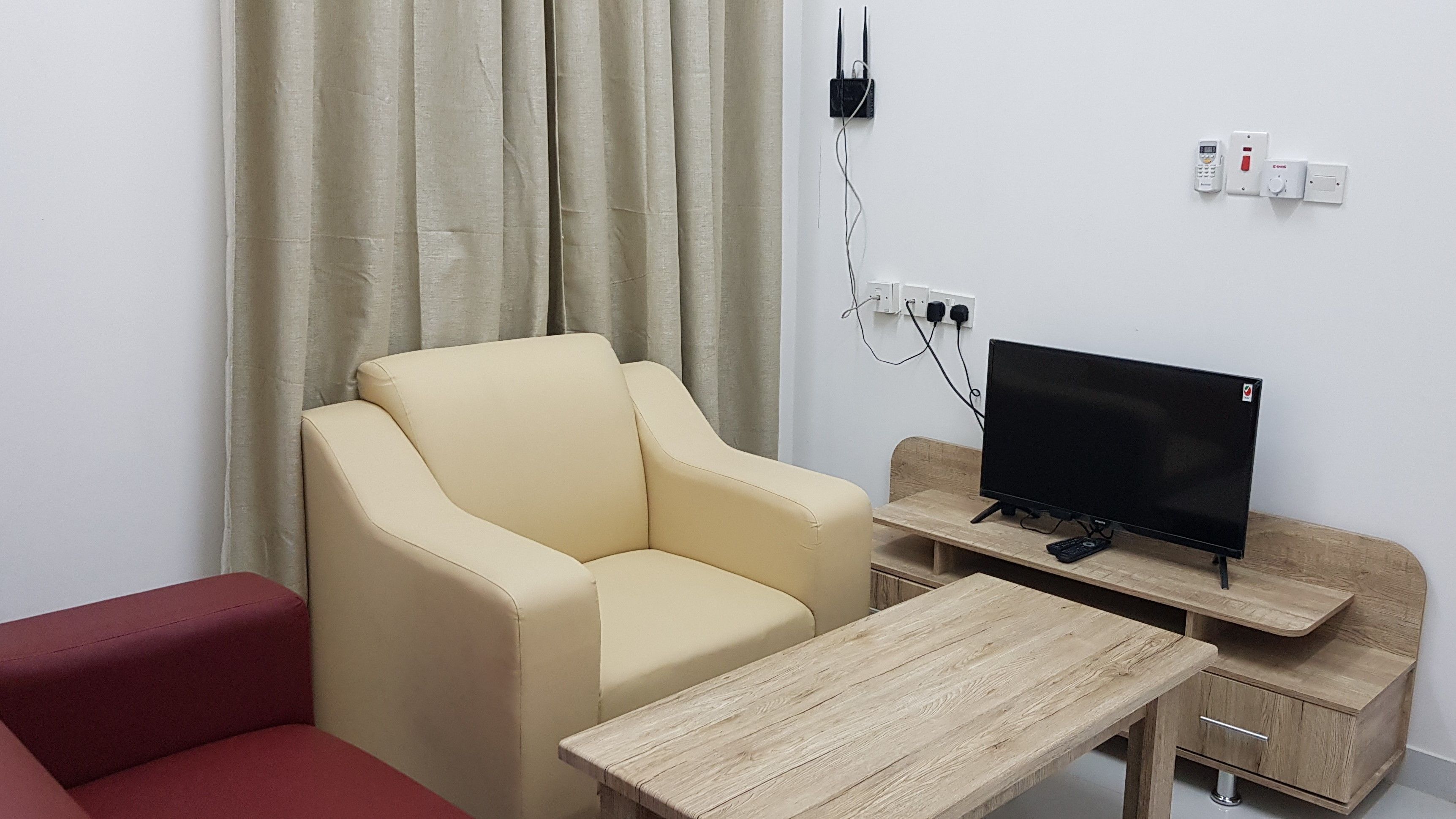 A living room with a beige sofa, a red armchair, a wooden coffee table, a TV on a wooden stand, and a wall-mounted power outlet with a phone and a switch.