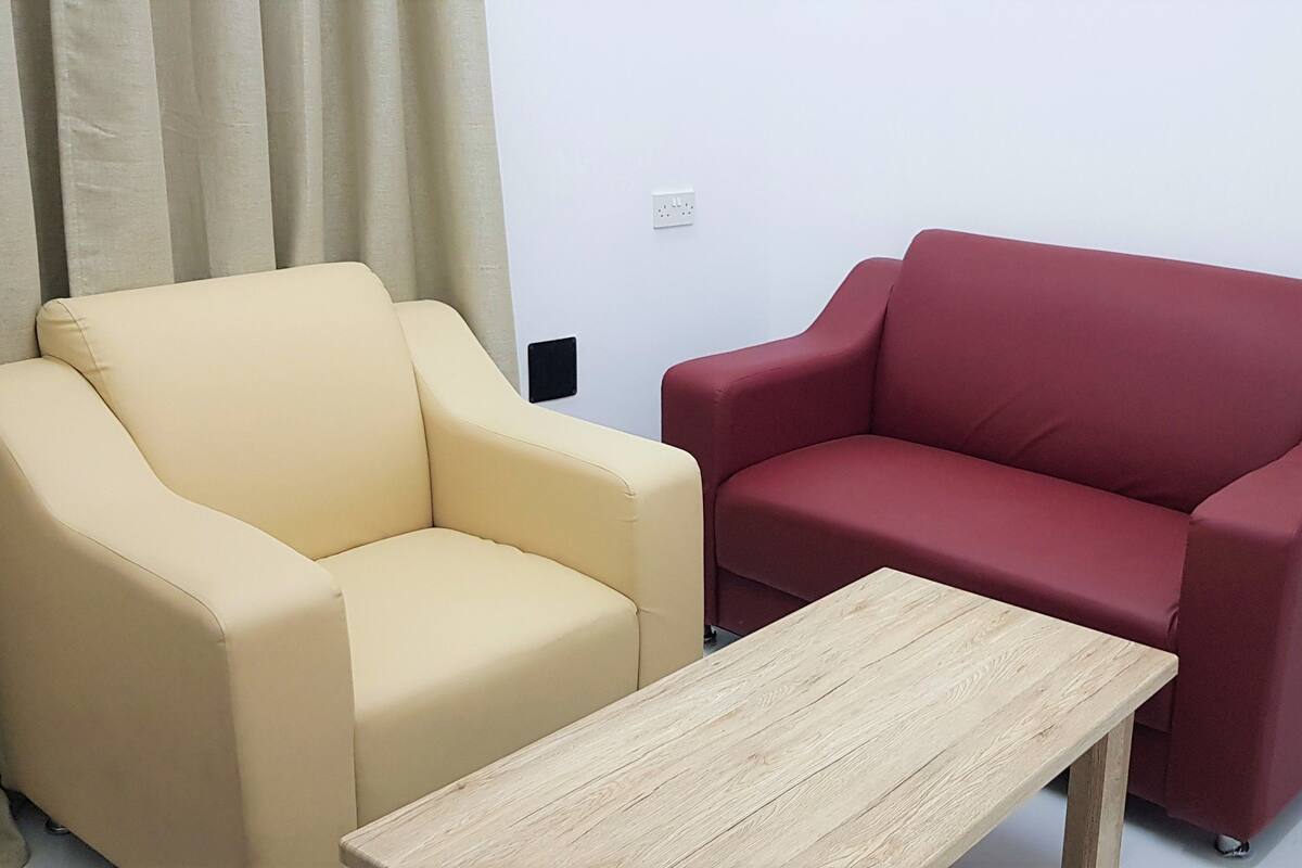 Una habitación con un sillón color beige y un sofá rojo, una mesa de centro de madera y una pared blanca con un enchufe.