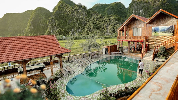 A resort with a swimming pool, wooden buildings, and a mountainous backdrop. - Windy Fields Villa (Nam Hoa Lu)