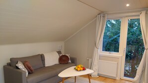 Een woonkamer met een grijze bank, een kleine witte tafel met fruit, en uitzicht op groen door een raam.