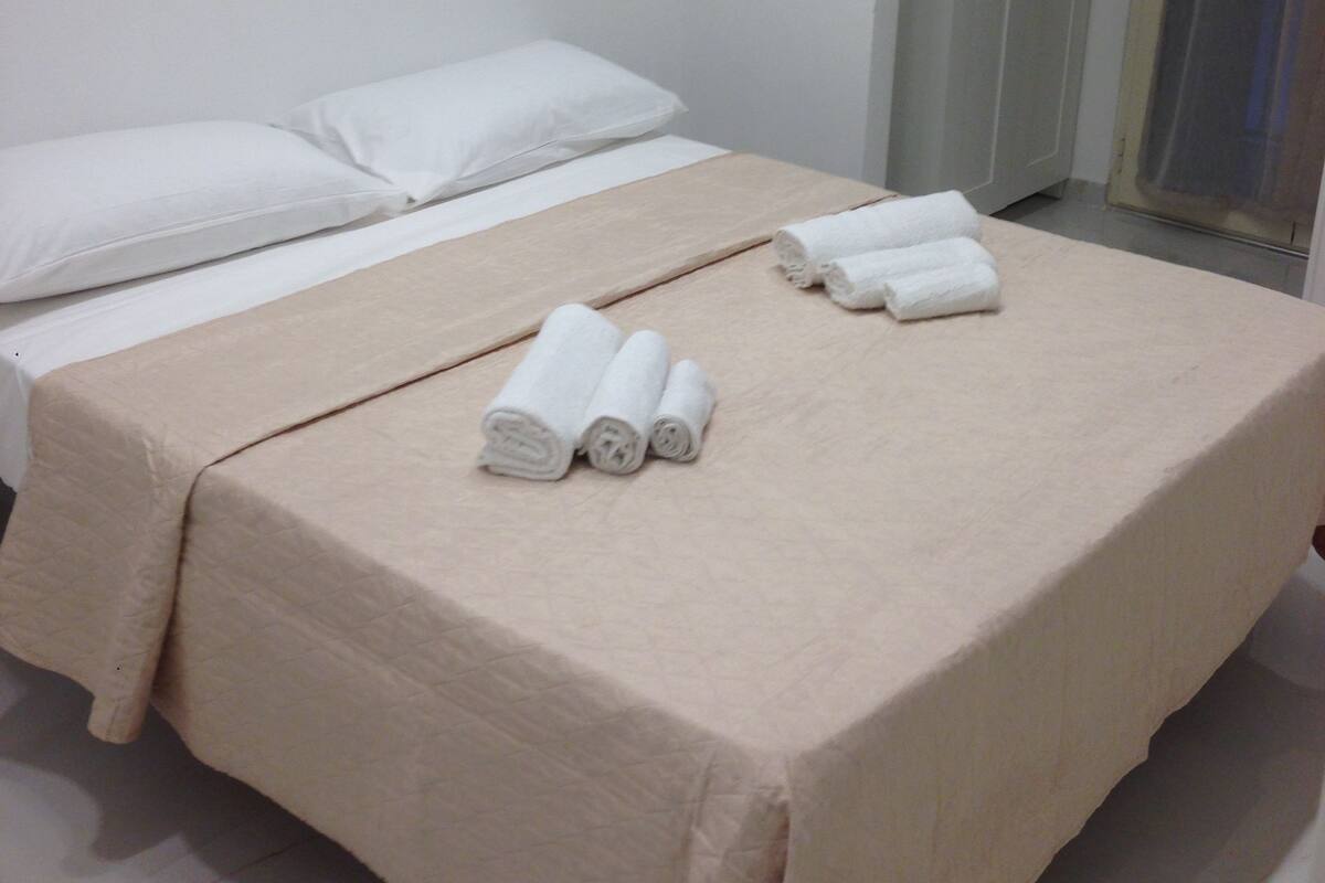 Una cama bien hecha con una colcha color beige y almohadas blancas, junto con toallas blancas enrolladas sobre la cama.