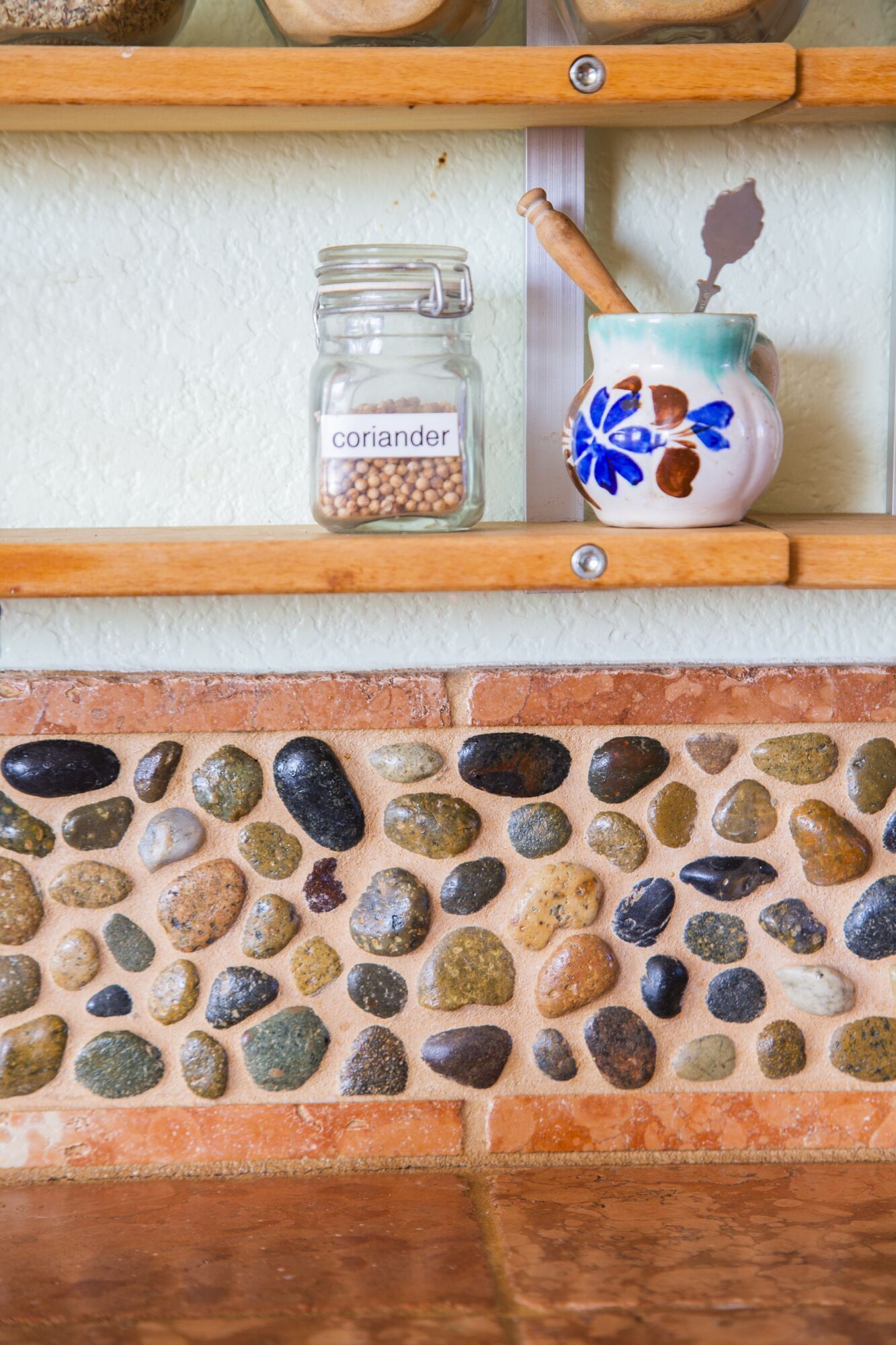 A shelf with a glass jar labeled "coriander" and a ceramic mug with a blue floral design. Below, a mosaic of variously colored stones is displayed.