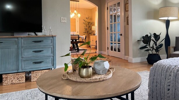 A living room with a TV, a blue cabinet, a round coffee table with decorative items, a lamp, and a doorway leading to another room. - Cody's Downtown Den - DOWNTOWN - New listing & Pets Welcomed! (Cody)