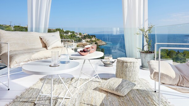 Un balcón moderno con un sofá blanco, mesas redondas y vistas al mar y un pueblo lejano.