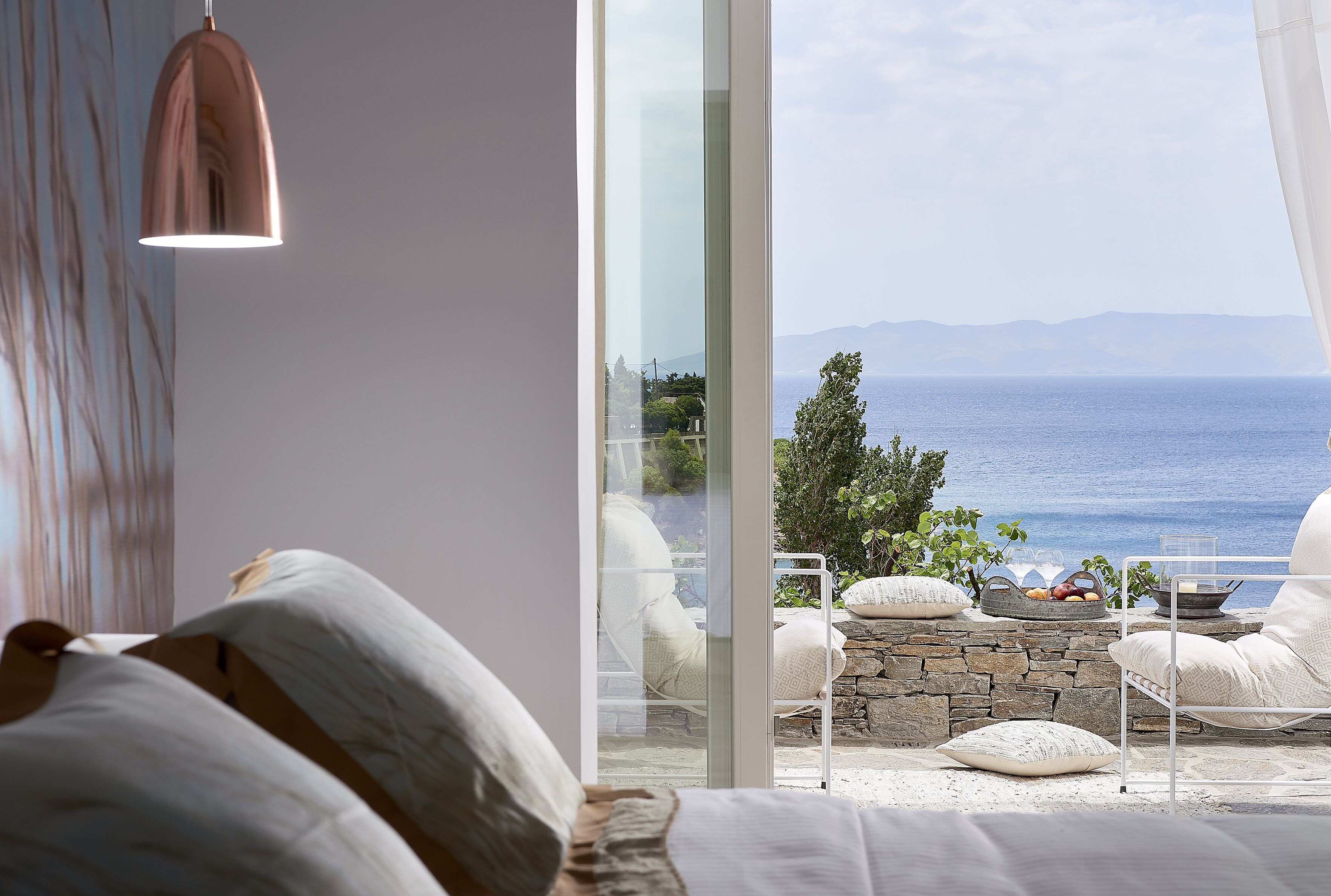 A bedroom with a large window overlooking the sea, a bed with white bedding, and a stone wall.