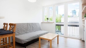 A modern living room with a grey sofa, wooden chairs, a small table, and large windows offering a view of the city. - Comfortable Studio in the Center | Parking and Balcony (Warszawa)