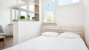 A minimalist bedroom with a bed, bedside tables, a chair, and a window with a city view. - Comfortable Studio in the Center | Parking and Balcony (Warszawa)